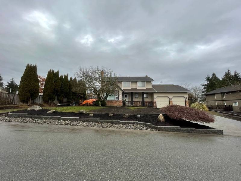 the street view showing the north side stack stone wall stepping down every 10 stones. there is 2"-6" river rock in front along the street and mulch on the other side. large boulders are placed in the mulch 